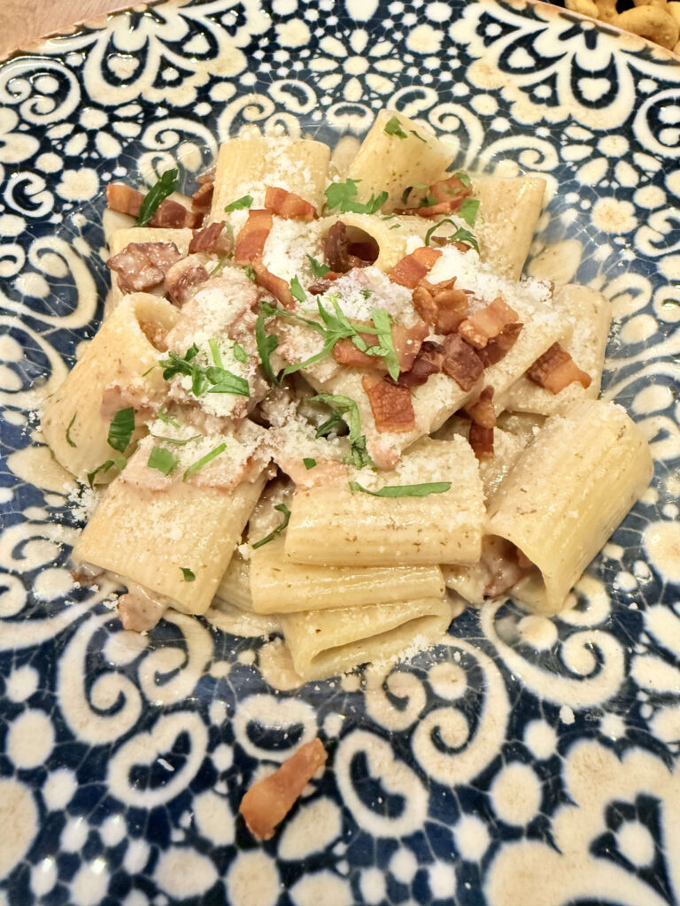 Rigatoni truffle carbonara topped with crispy bacon, grated cheese, and herbs served on a blue and white patterned plate