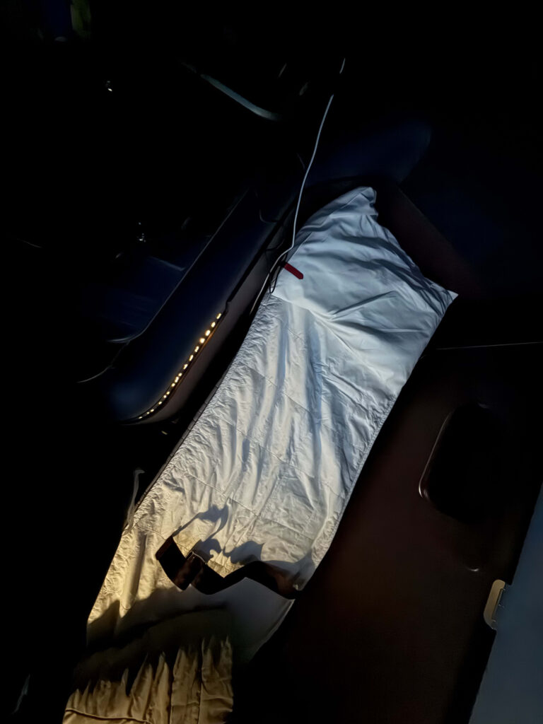 Full-flat airline seat set up as a bed with white airweave bedding in a dimly lit cabin