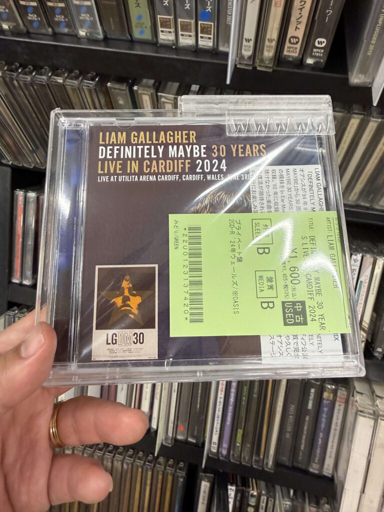 CD棚を背景に、リアム・ギャラガーの『Definitely Maybe 30 Years Live In Cardiff 2024』というCDを手に持っている様子。緑色の値札シールが貼られている。