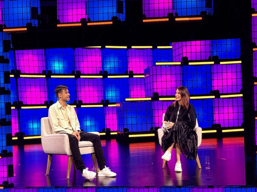 A man and a woman sitting in white armchairs on a stage with a background of stacked blue and pink illuminated cubes during the Lovable session