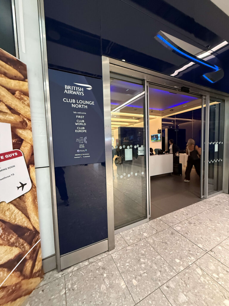 Entrance to the British Airways Club Lounge North at Heathrow Airport featuring a dark blue sign and glass automatic doors.