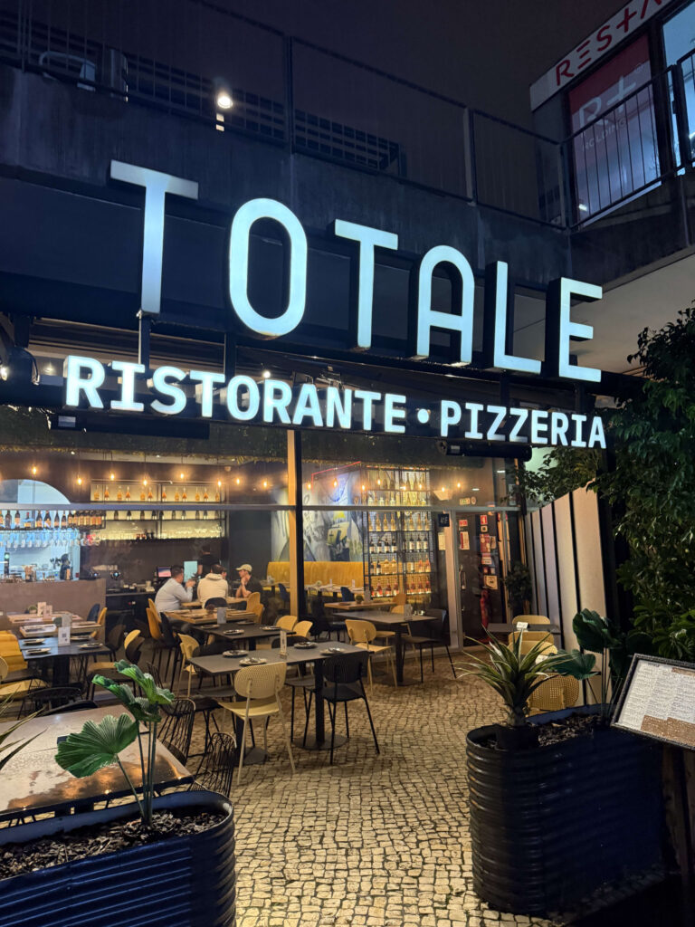Exterior view of TOTALE RISTORANTE PIZZERIA at night, featuring large illuminated signage, glass windows, and outdoor seating on cobblestones.