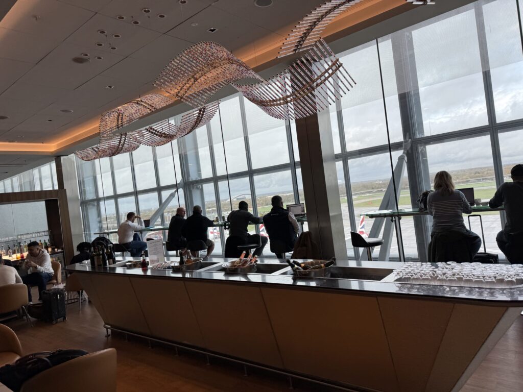 Travelers working on laptops at a window counter in an airport lounge with a large wave-shaped light fixture and bar area