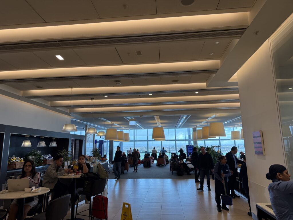 Interior view of the British Airways North Lounge featuring high ceilings, large pendant lights, and travelers relaxing at tables and armchairs.