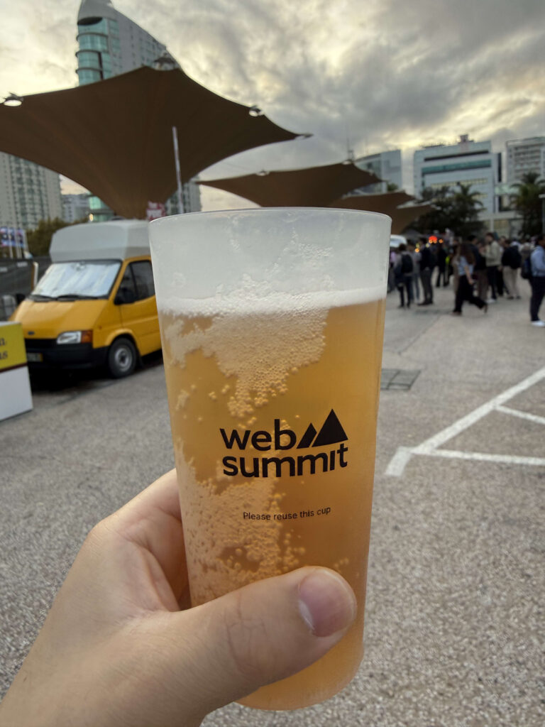 Web Summitのロゴが入ったリユースカップに入ったビールを手で持っている様子。背景には黄色いバンとイベント会場の人混みが見える。
