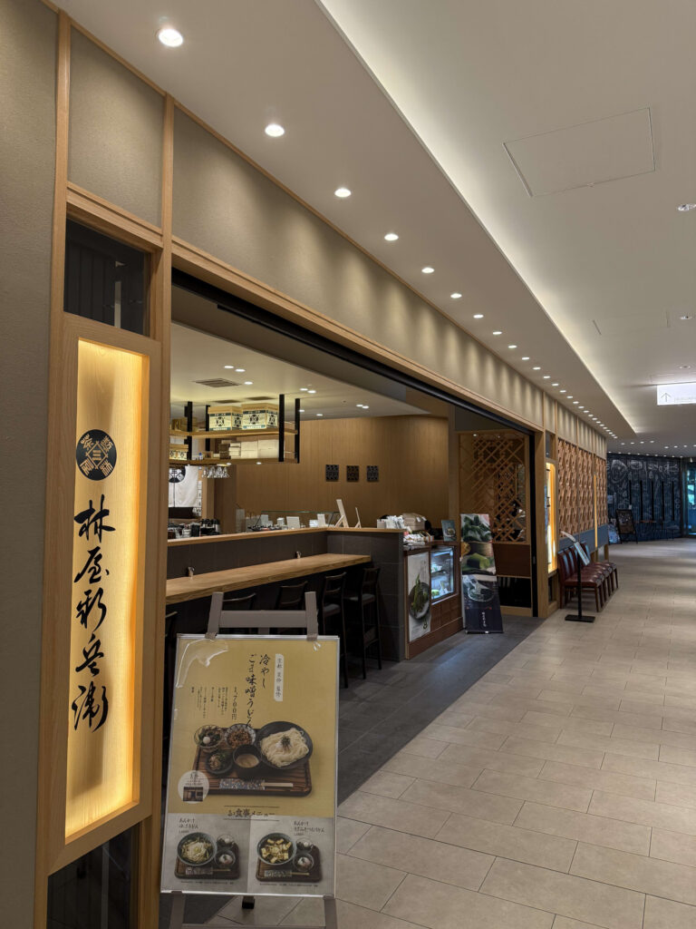 東京ミッドタウン日比谷にある「日比谷 林屋新兵衛」の木目調の店舗外観と、手前に置かれた冷やしうどんのメニュー看板