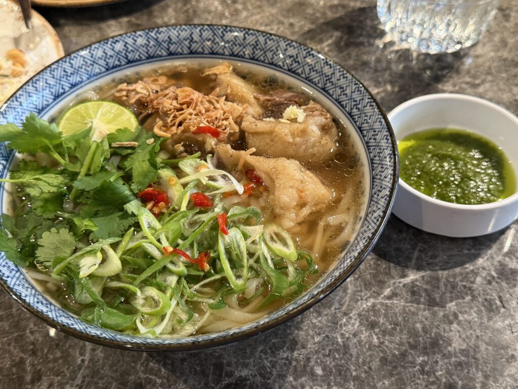 Oxtail Pho in a blue and white bowl topped with cilantro, green onions, lime, and chili, served with a side of green sauce