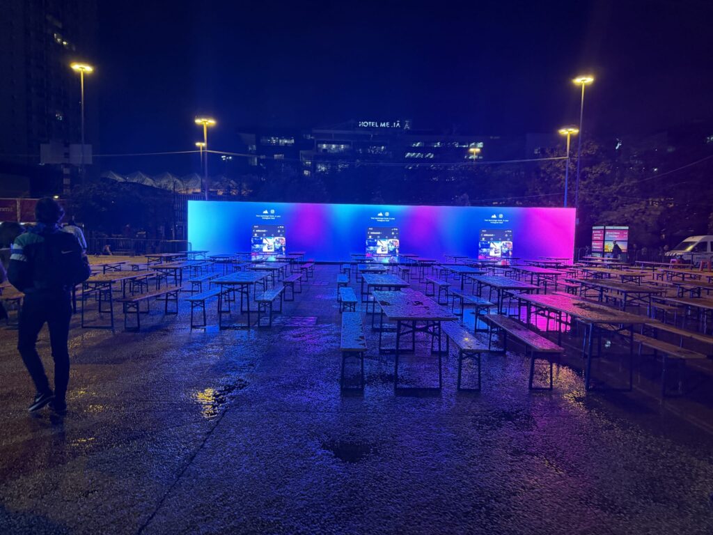Night view of the Web Summit outdoor venue with empty tables and benches. Bright blue and magenta screens reflect on the wet pavement as a person walks away.