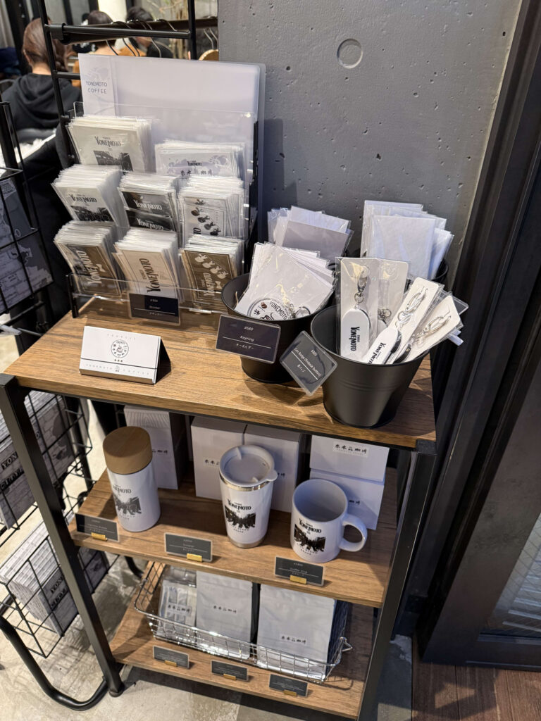 Wooden display shelf featuring Yonemoto Coffee original merchandise, including drip bags, keychains, tumblers, and mugs against a concrete wall.