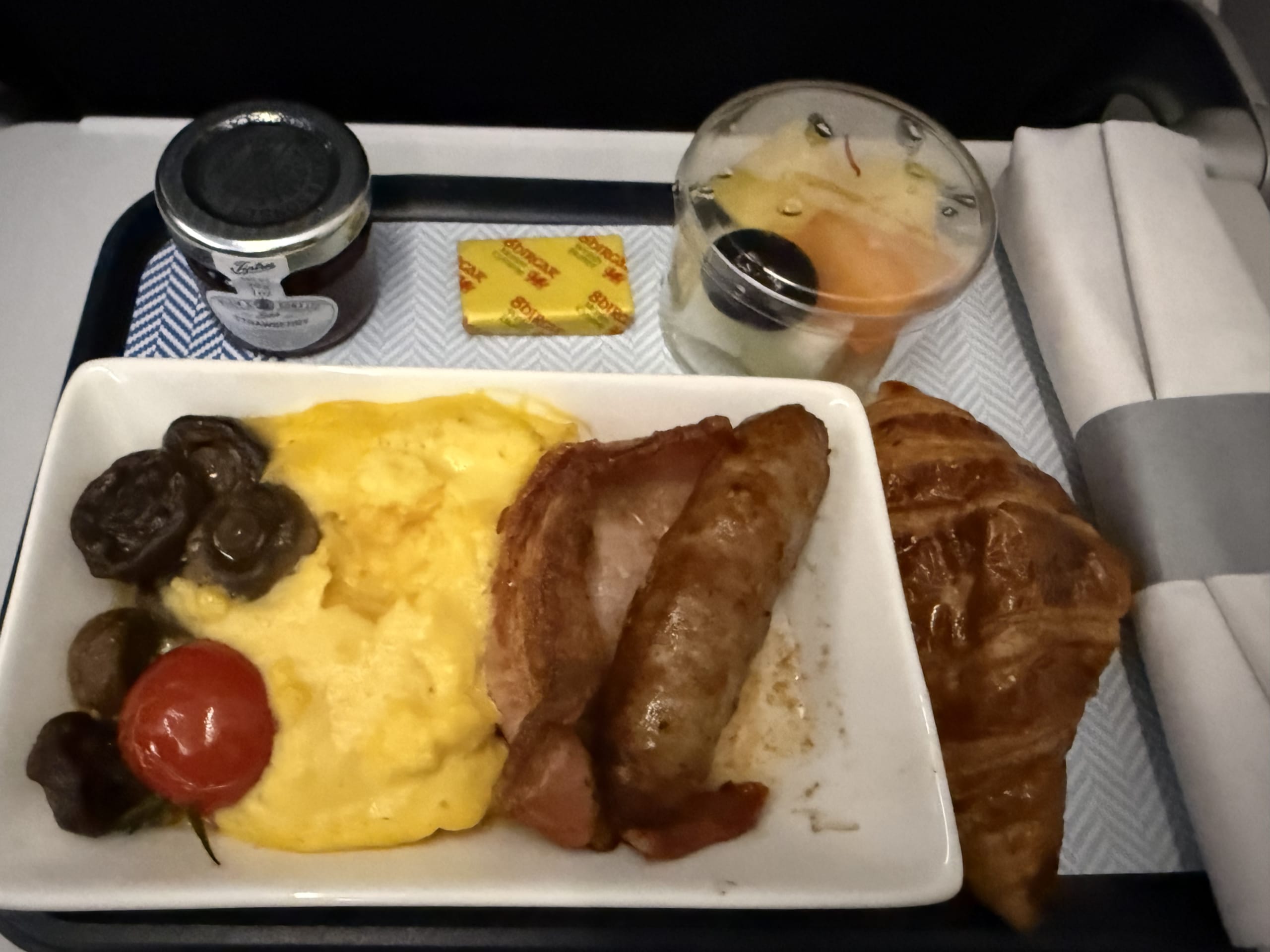 Airline meal tray featuring 'The Original Full British' breakfast with scrambled eggs, bacon, sausage, mushrooms, tomato, a croissant, and fruit cup.