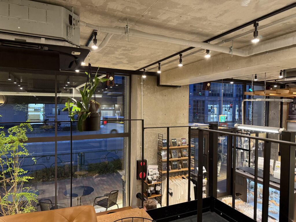 Interior view of a modern cafe with high concrete ceilings, exposed pipes, large windows overlooking a street, and hanging plants
