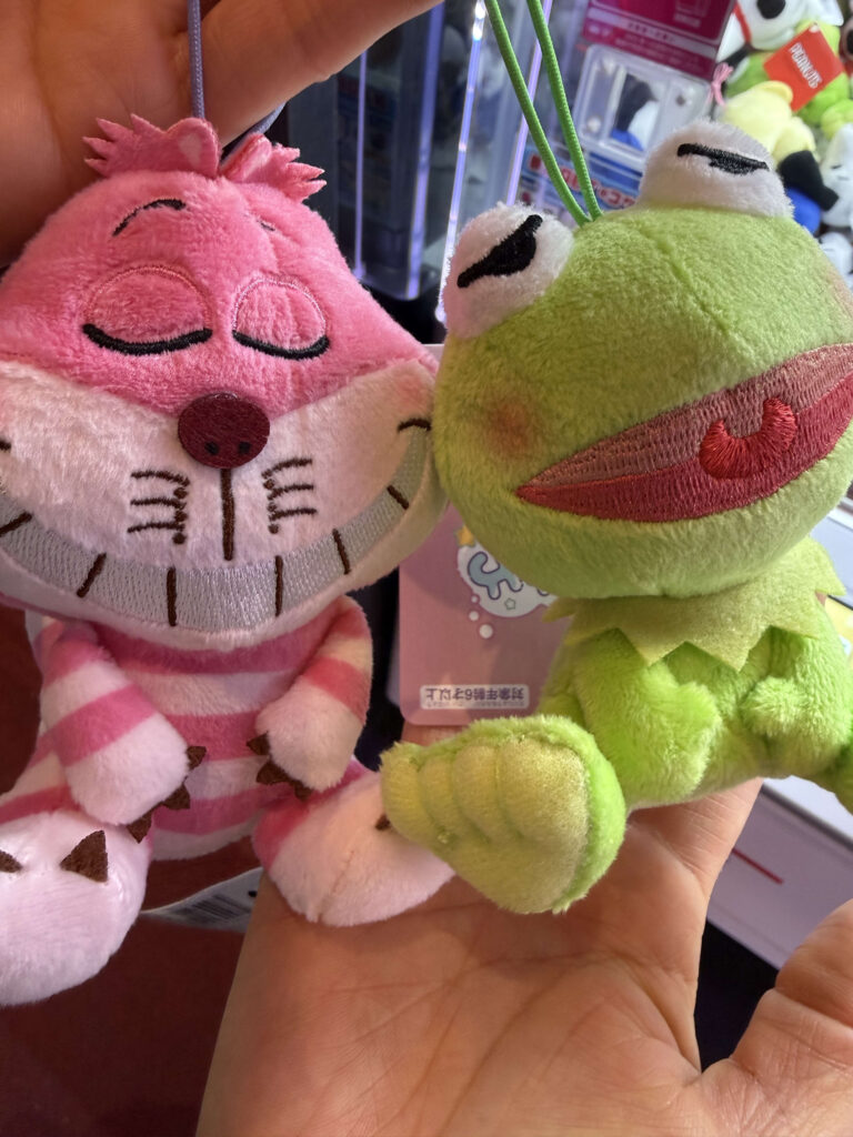 Pink Cheshire Cat and green Kermit the Frog plush toys held in a hand
