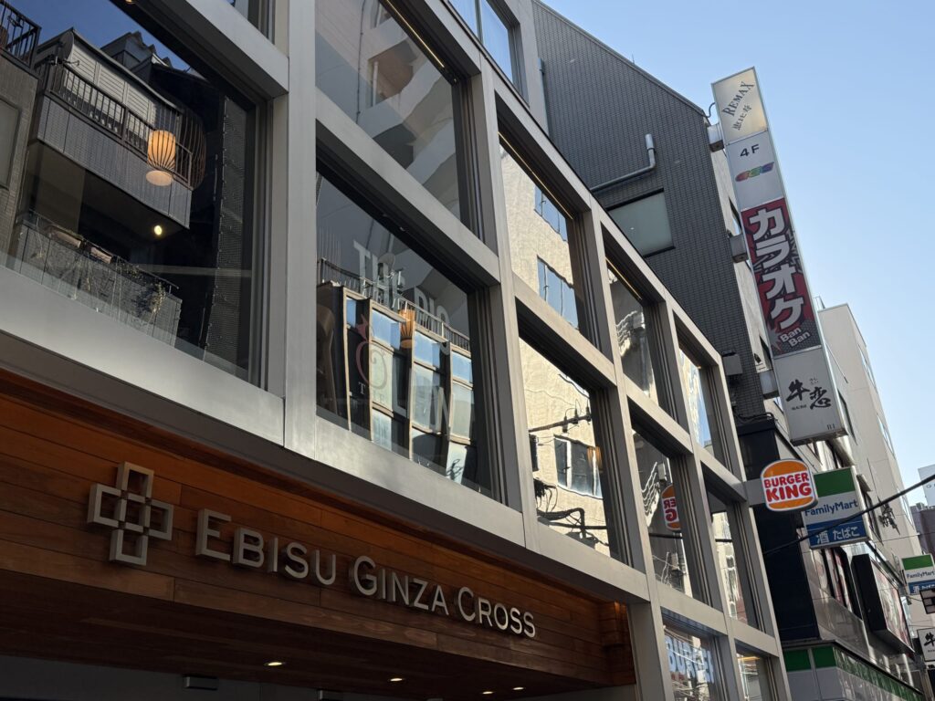 青空の下、大きなガラス窓が特徴的な商業ビル「EBISU GINZA CROSS」の外観。木製のビル名サインと、バーガーキングやカラオケ店、牛角などのテナント看板が見える