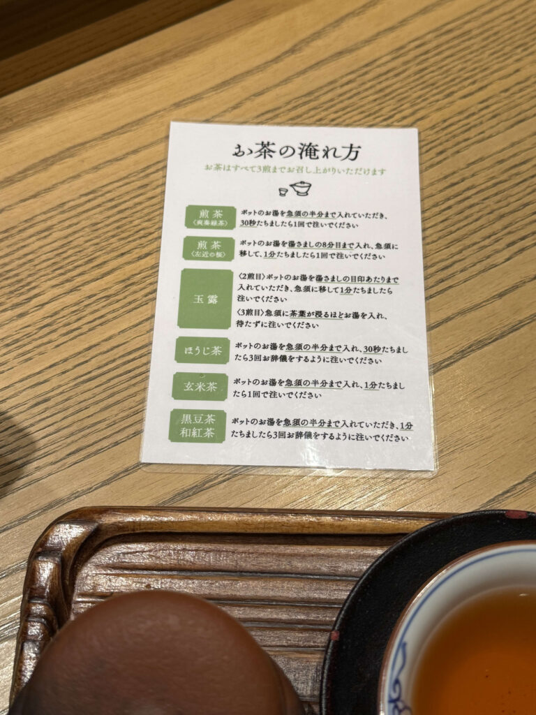 A tea brewing instruction card on a wooden table listing methods for Sencha, Gyokuro, and Hojicha, with a tea treat in the foreground