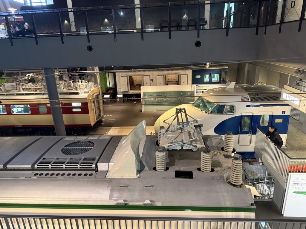Overhead view of the Railway Museum Vehicle Station, featuring a close-up of the roof and pantograph of a 0 Series Shinkansen in the foreground and a red and cream limited express train in the background.
