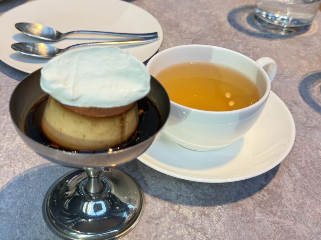 Classic custard pudding topped with whipped cream served in a silver footed bowl next to a cup of tea