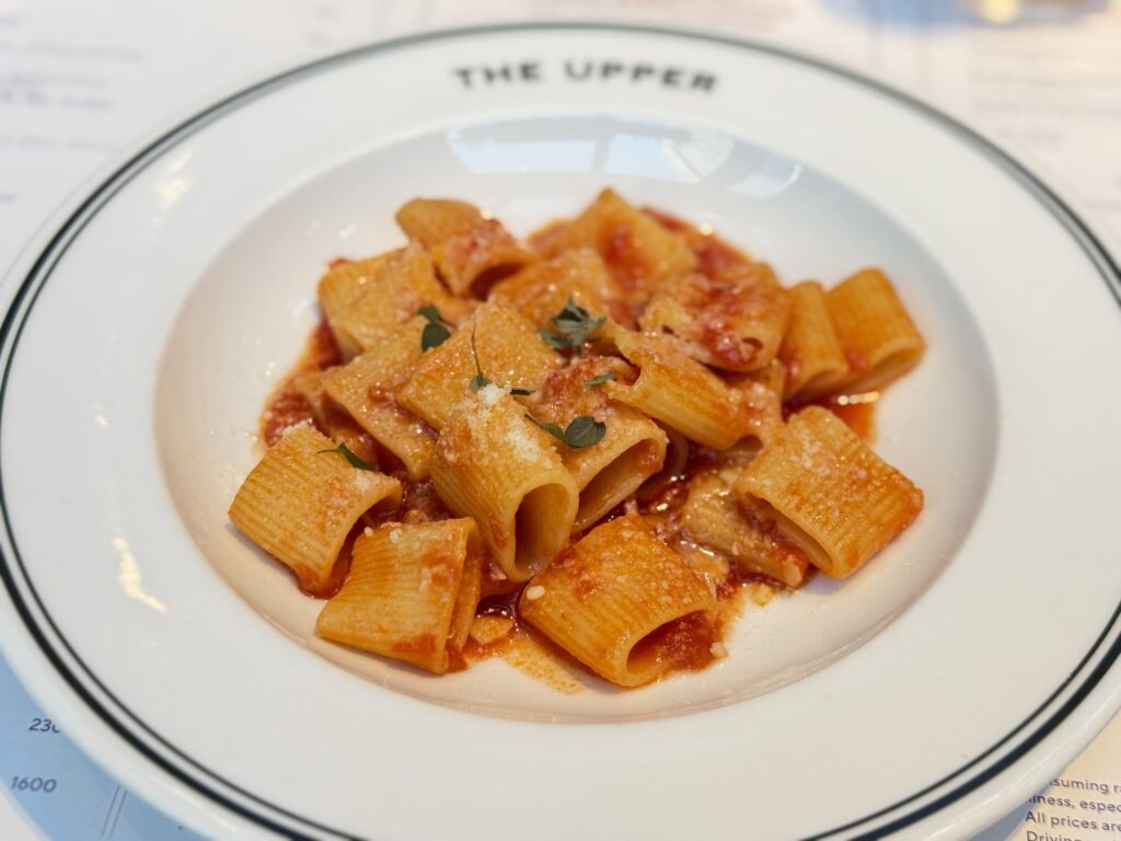 Short pasta with tomato sauce topped with cheese and herbs served on a white plate with THE UPPER text