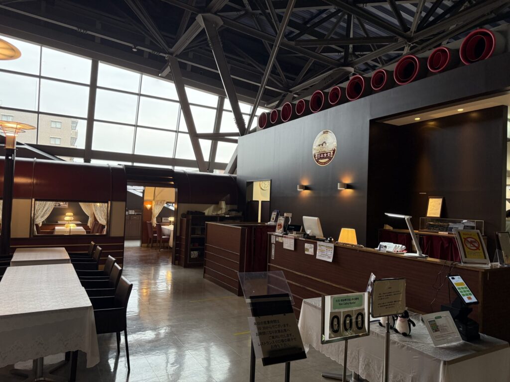 鉄道博物館にあるトレインレストラン日本食堂の入り口。左手には食堂車を模した客席、中央には受付カウンターと順番待ちシステム、背景には高い天井と大きな窓が見える。