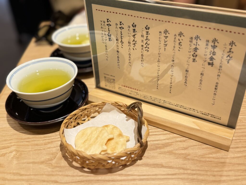 木製のテーブルの上に置かれた甘味処のメニュー表、緑茶が入った湯呑み、竹籠に入った豚の形のお茶請け（せんべい）