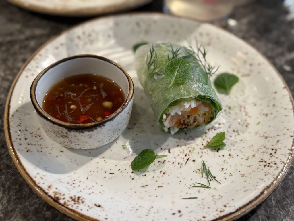 A fresh spring roll on a speckled ceramic plate garnished with herbs, served with a small bowl of dipping sauce