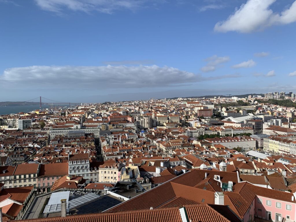 Panoramic view of Lisbon's cityscape with dense red roofs, white buildings, and a bridge in the distance under a blue sky