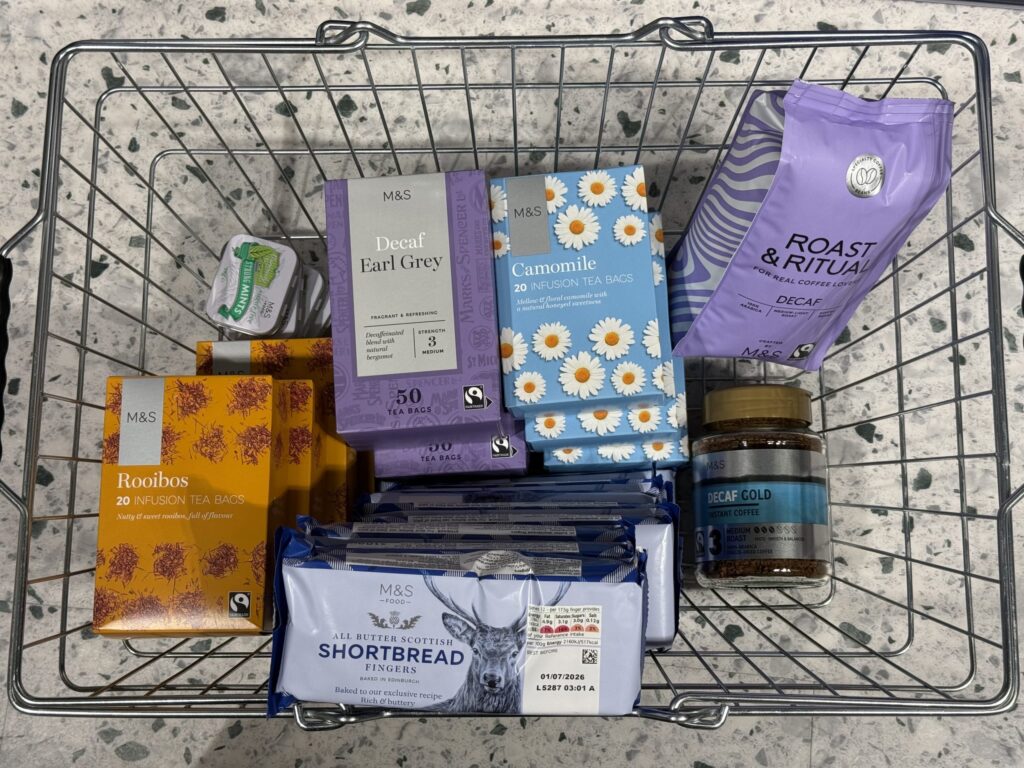 A wire shopping basket filled with Marks & Spencer products including Rooibos tea, Decaf Earl Grey, Camomile tea, coffee beans, instant coffee, shortbread fingers, and mints
