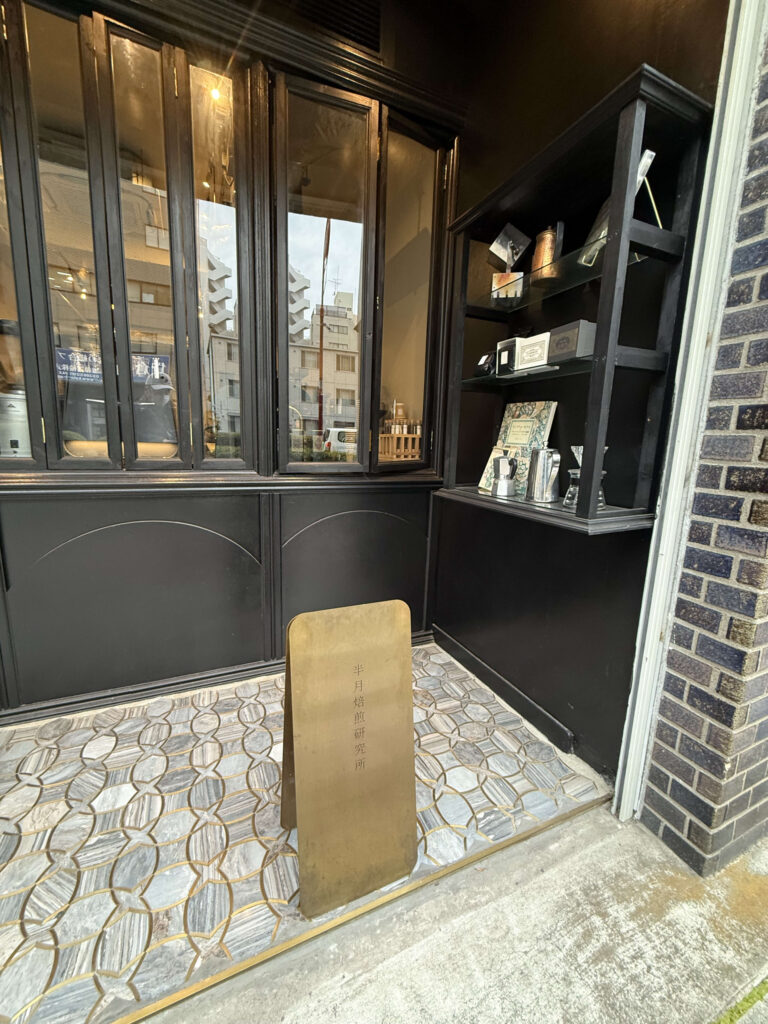 Storefront entrance featuring black-framed glass folding doors, a geometric mosaic tile floor, and a brass standing sign reading 'Hangetsu Baisen Kenkyujo'.