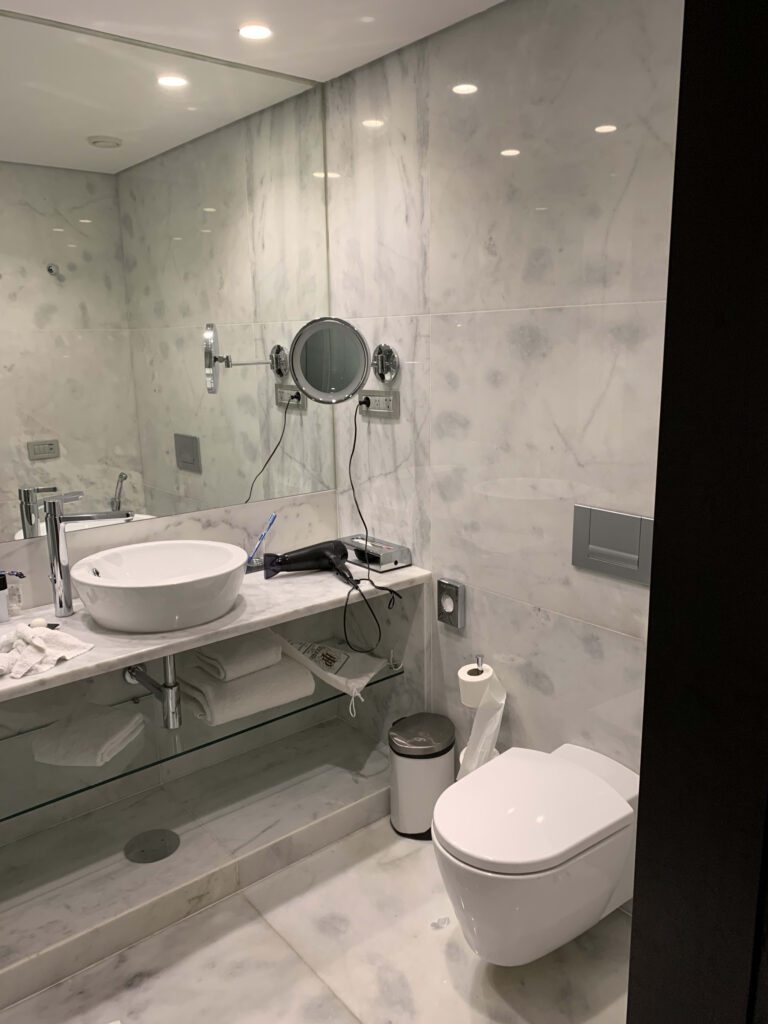 Modern bathroom featuring white marble-effect tiles, a round vessel sink, wall-mounted magnifying mirror, hairdryer, and a white toilet.