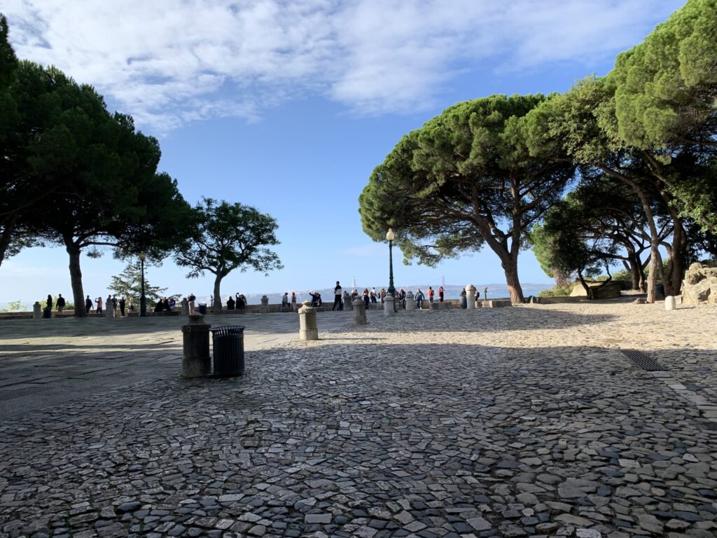 Cobblestone plaza at a castle viewpoint (Miradouro) with large pine trees and tourists looking at the view