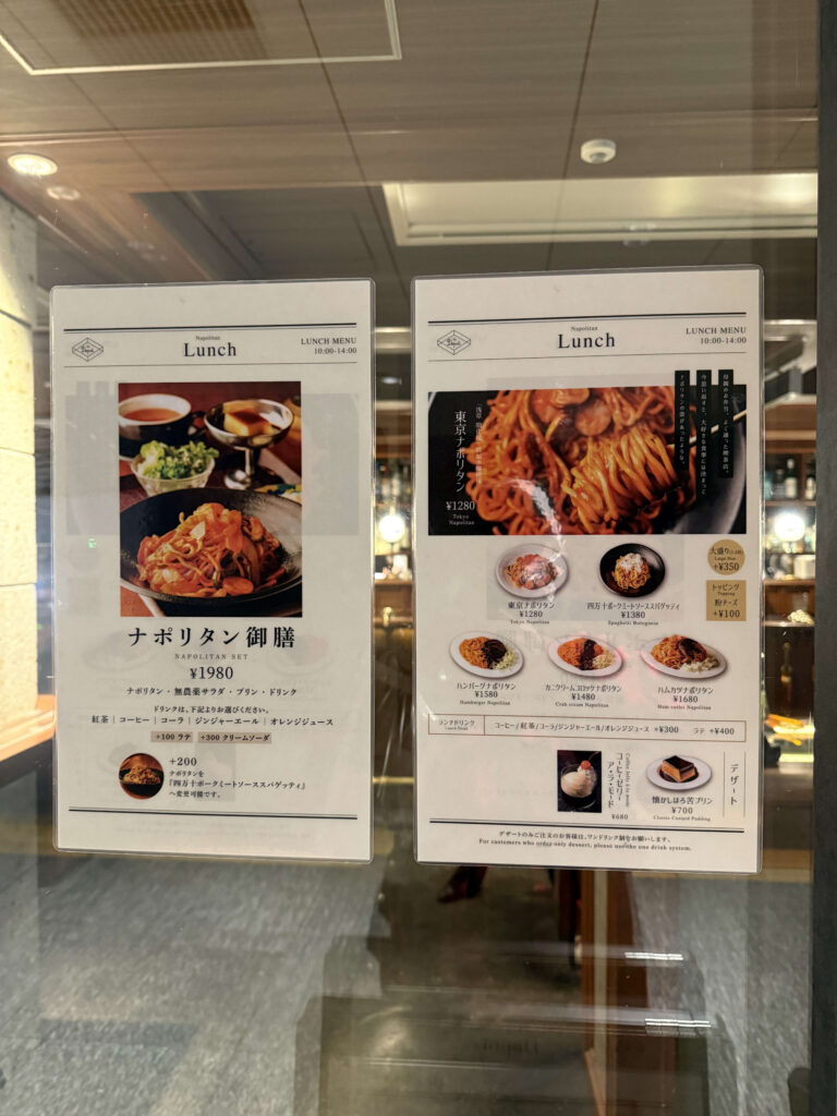 ガラス越しに掲示された飲食店のランチメニュー表。ナポリタン御膳や東京ナポリタン、デザートの写真と価格が記載されている。