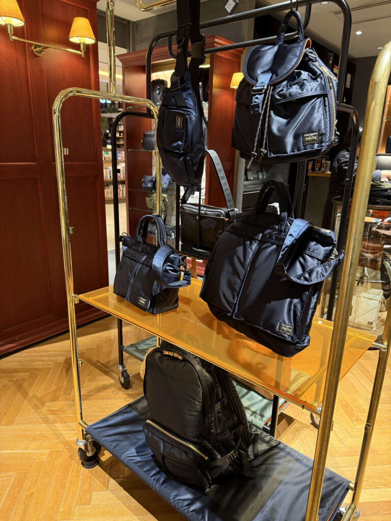 Store display at Marunouchi KURA CHIKA featuring navy Porter Tanker series bags hanging on a gold rack, with a large XL backpack resting on the bottom shelf
