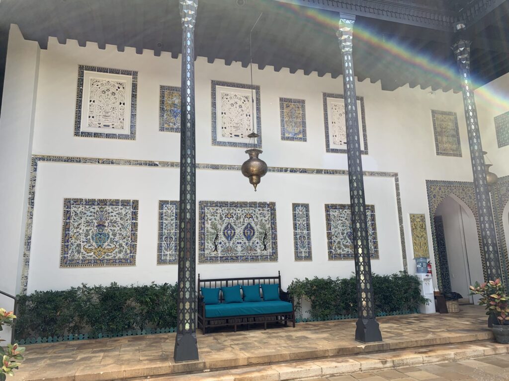 Full view of a courtyard featuring white walls decorated with geometric tile panels, a bench with turquoise cushions, and tall decorative columns