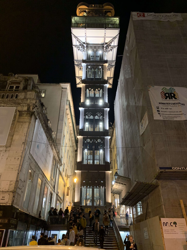 The illuminated Santa Justa Lift tower in Lisbon at night, highlighting its ironwork against the dark sky with people gathered on the stairs below