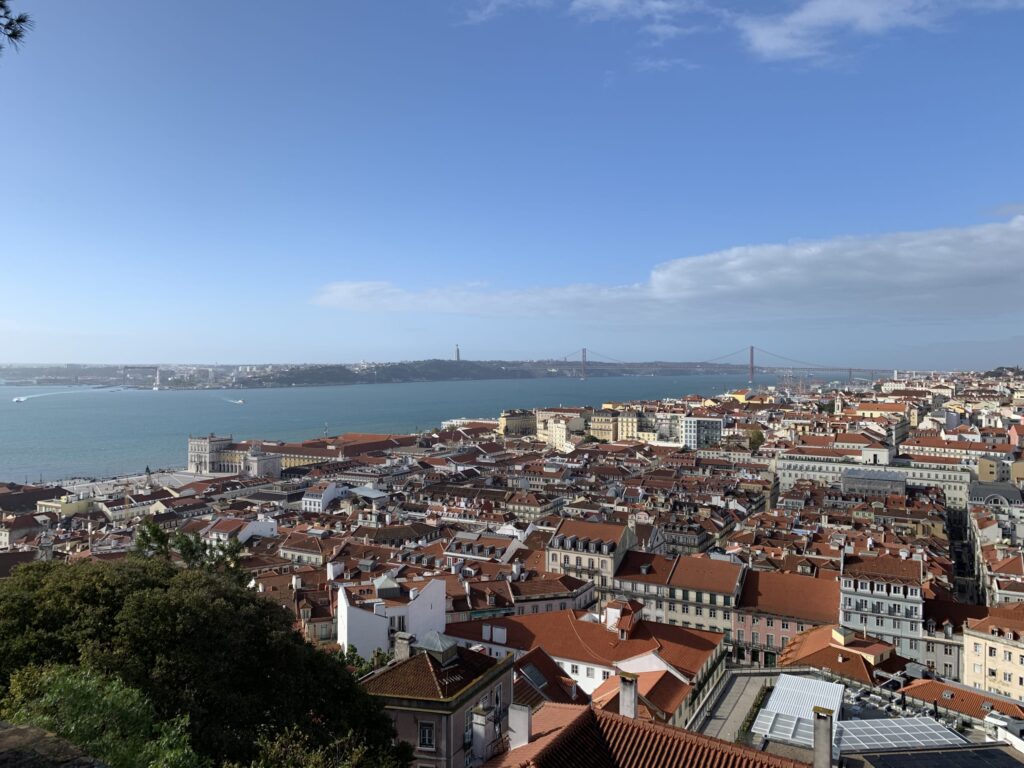 View of Lisbon's orange rooftops, the wide Tagus River, and the 25th of April Bridge under a blue sky