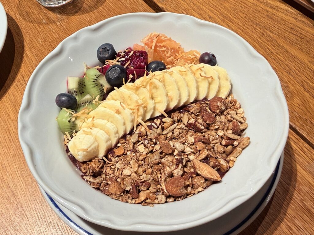 Acai bowl served in a white scalloped bowl, topped with sliced bananas, kiwi, blueberries, citrus segments, and granola on a wooden table.
