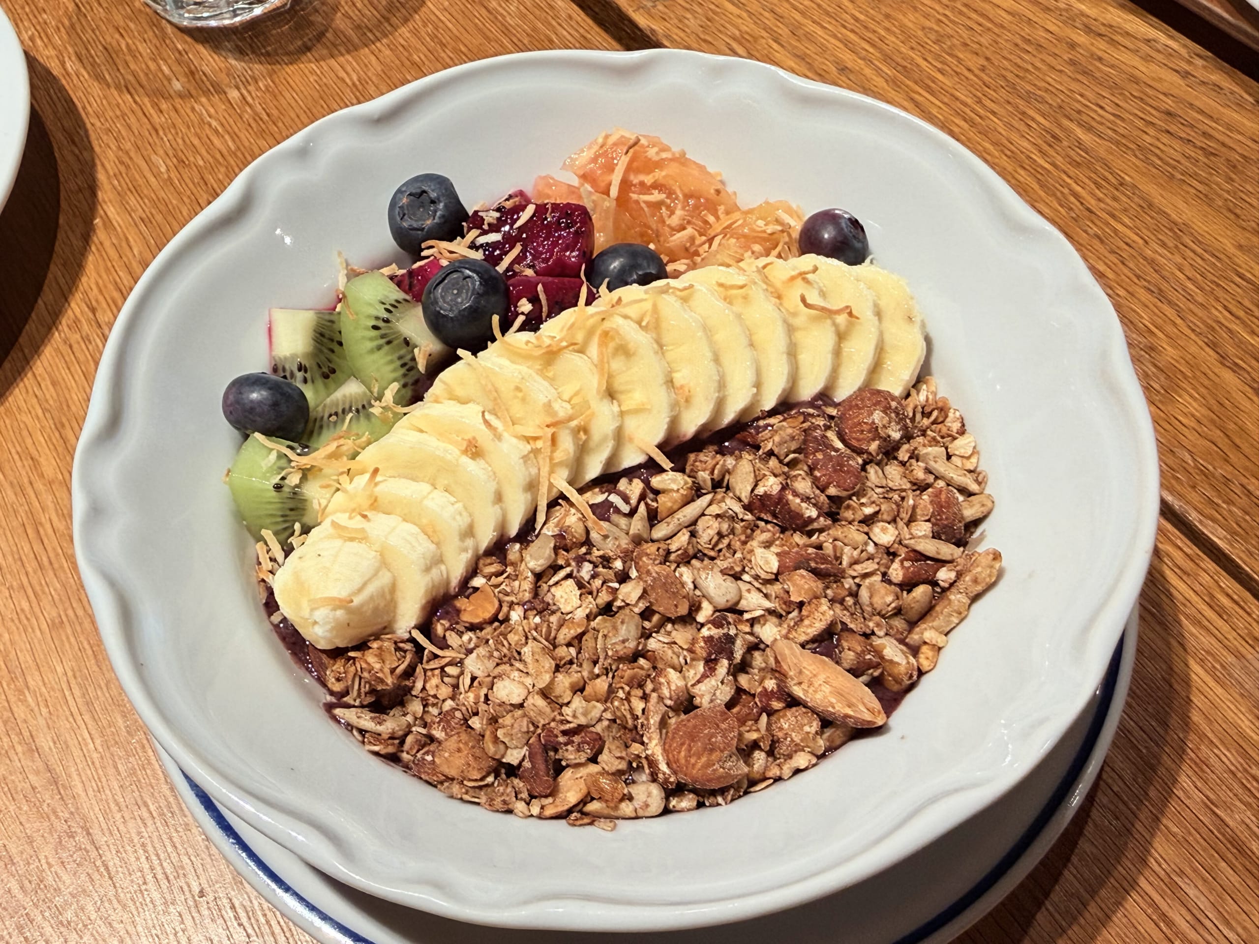 Acai bowl served in a white scalloped bowl, topped with sliced bananas, kiwi, blueberries, citrus segments, and granola on a wooden table.
