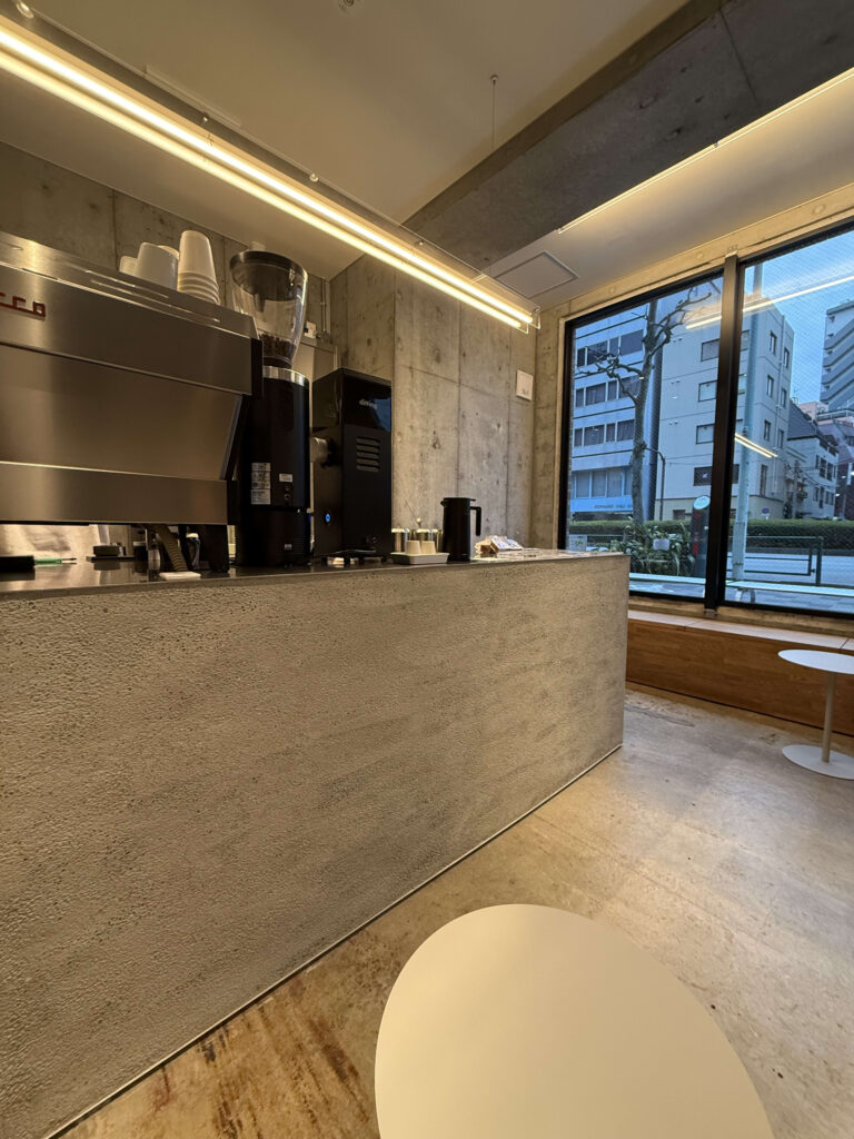 A stylish cafe interior featuring a concrete mortar counter with an espresso machine and a large window overlooking a street