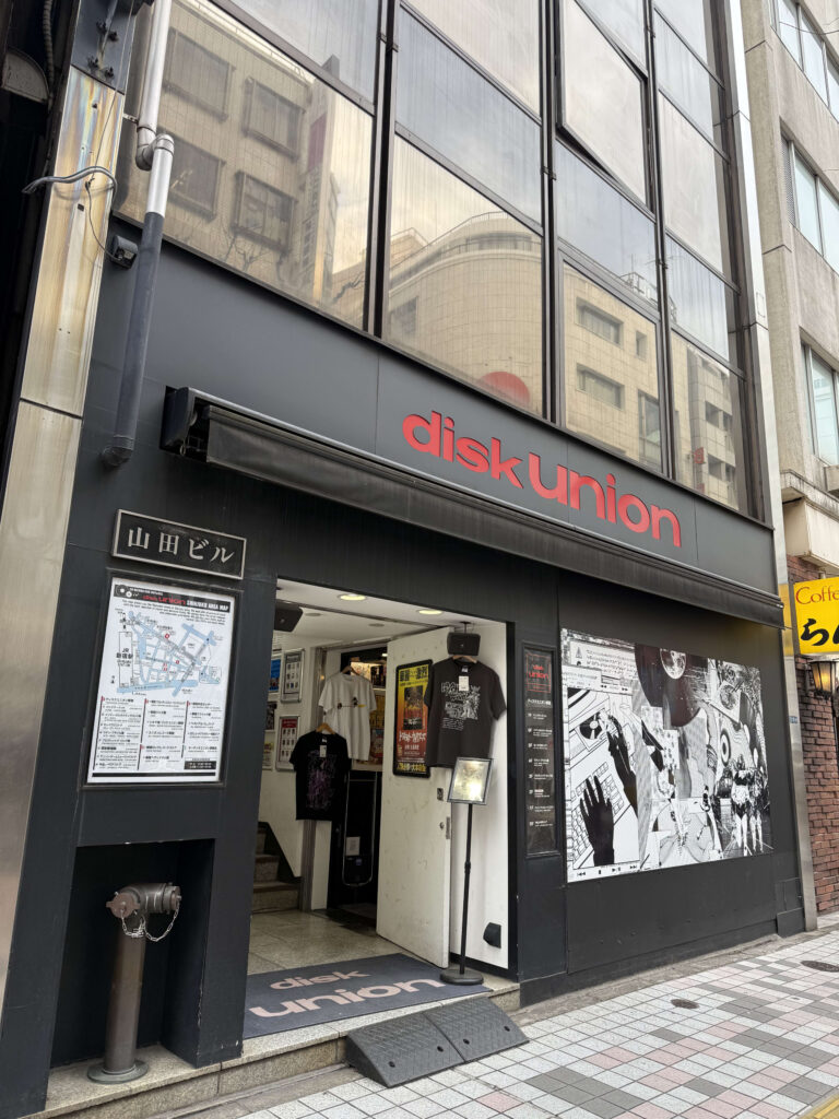 Exterior of the Disk Union store in Shinjuku's Yamada Building, featuring a black facade with a large red logo and manga-style wall art.