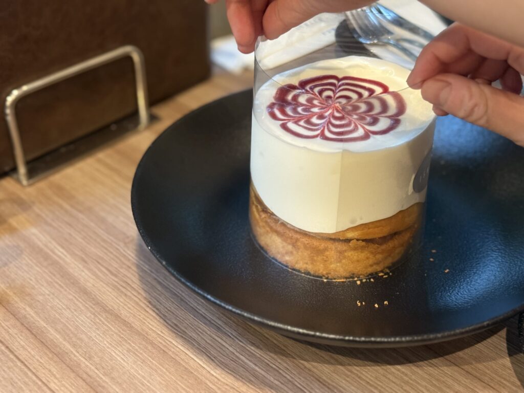 Hands lifting a clear plastic sheet surrounding a thick pancake topped with abundant white cream and a red berry swirl pattern on a black plate