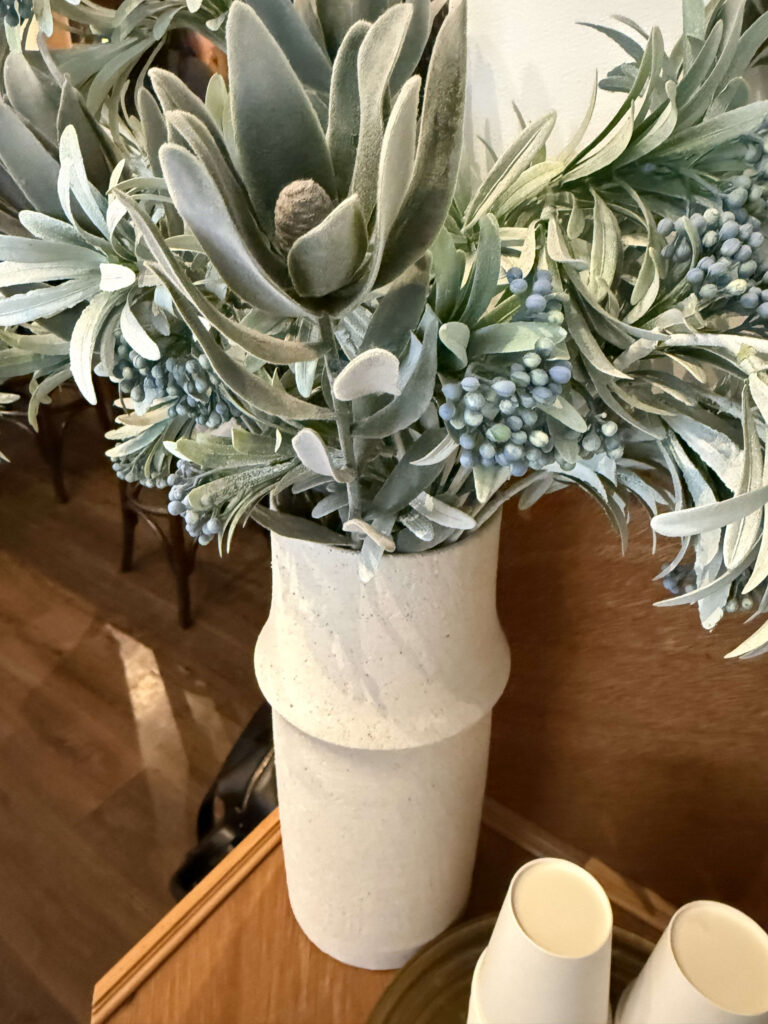 Dusty green plant arrangement with blue berries in a textured white vase on a wooden table