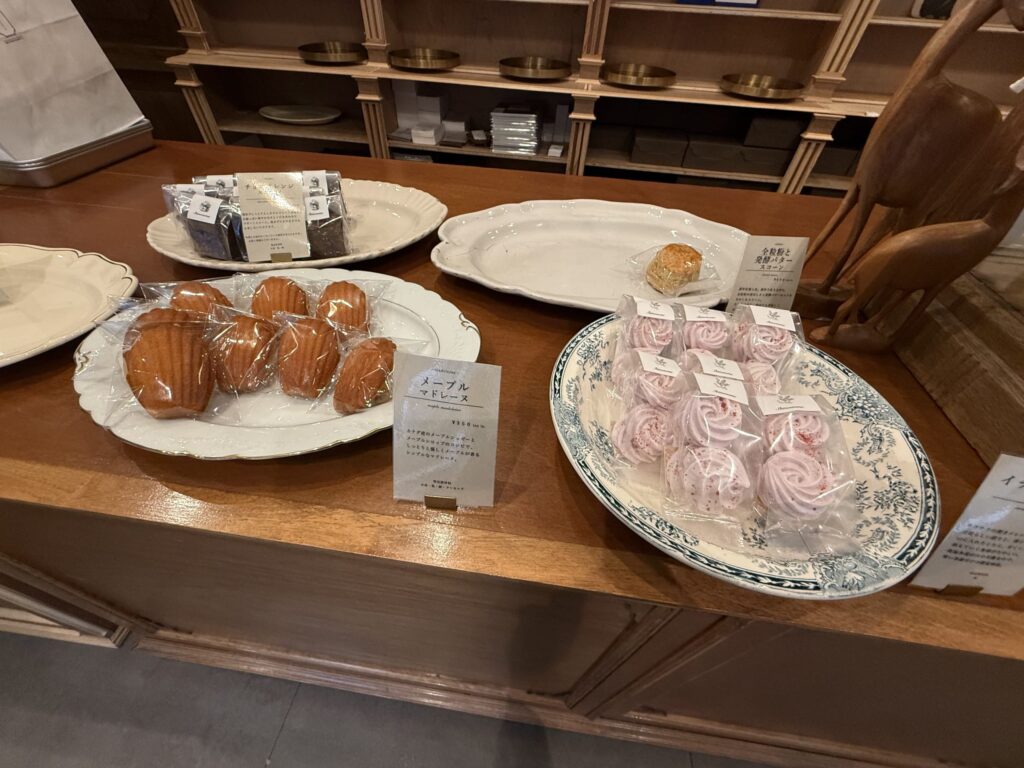 木製のカウンターテーブルに並べられたメープルマドレーヌ、ピンク色のメレンゲ、全粒粉スコーンなどの焼き菓子