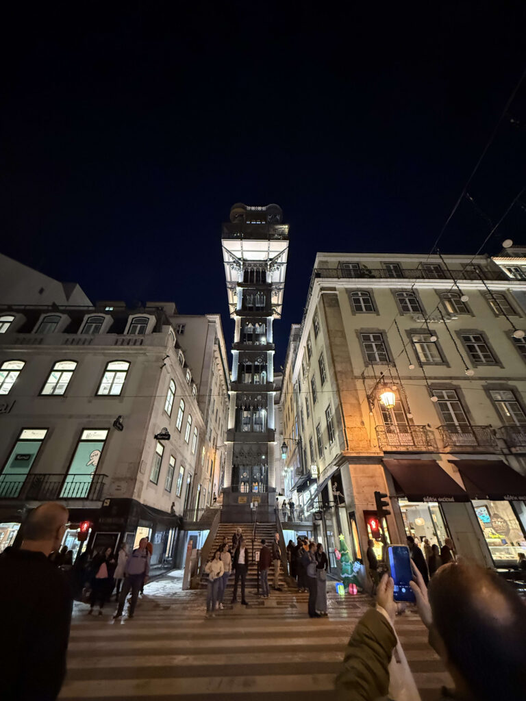 Tourists looking up and taking photos of the illuminated Santa Justa Lift at night with their smartphones