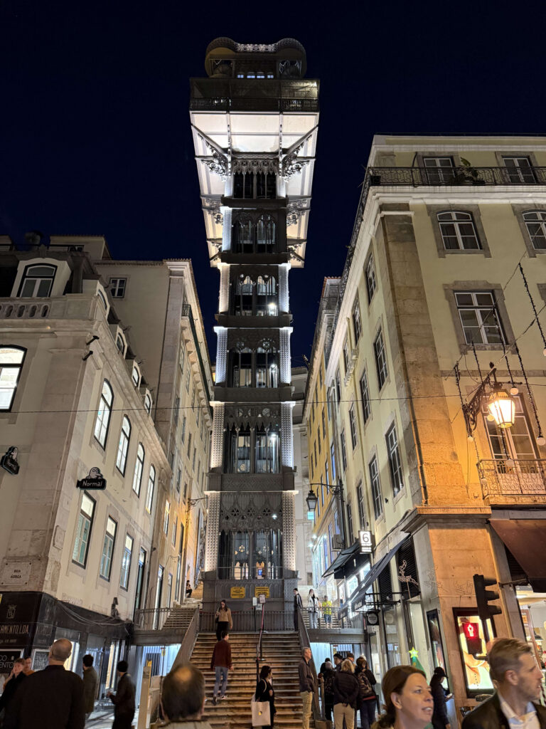 Low-angle night view of the illuminated Santa Justa Lift in Lisbon, with people walking on the stairs in the foreground