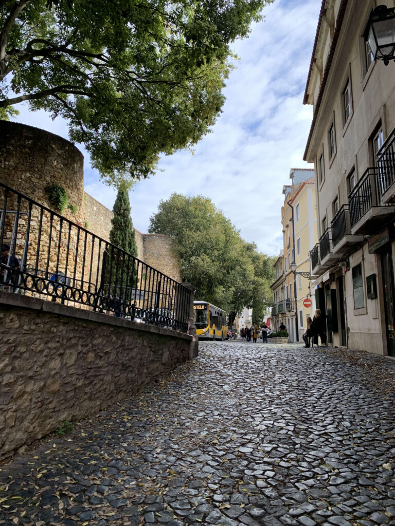 A cobblestone street in Lisbon along a historic stone wall, featuring a yellow bus in the distance and fallen leaves