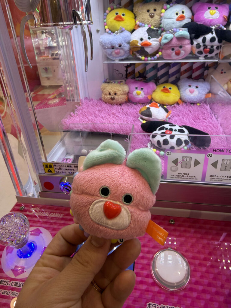 Hand holding a small pink mojojojo bear plush toy with a red heart nose in front of a claw machine