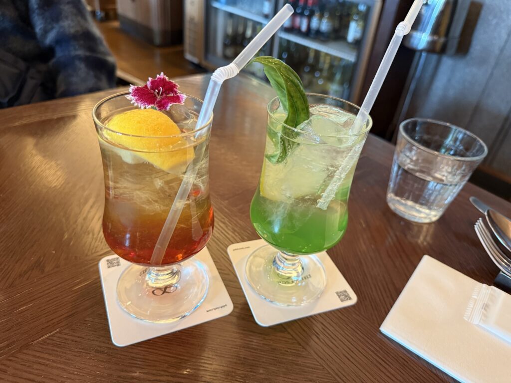 Amber-colored Pear Jasmine Blossom drink with flower garnish and green Basil and Kiwi Squash on a wooden table