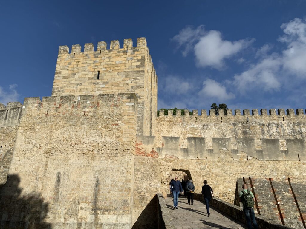 A tall stone castle tower and fortified walls under a blue sky, with tourists walking along a stone path toward the entrance of the inner citadel