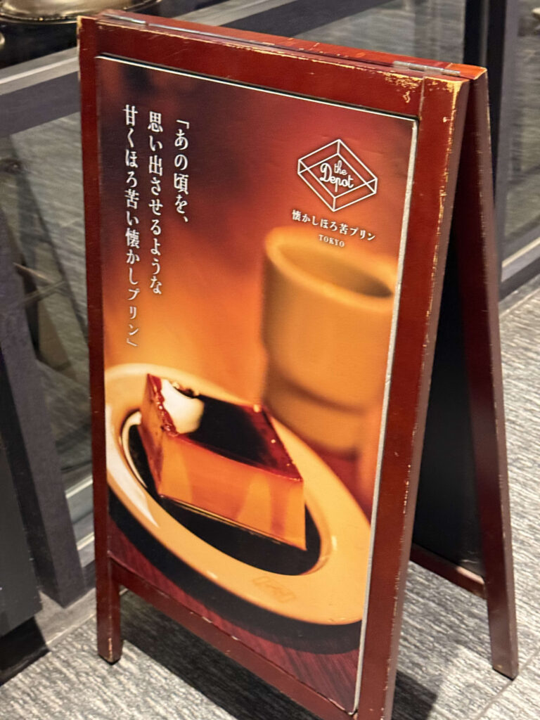 Red wooden signboard for 'The Depot' featuring a photo of a square pudding and Japanese text about nostalgic bittersweet pudding.