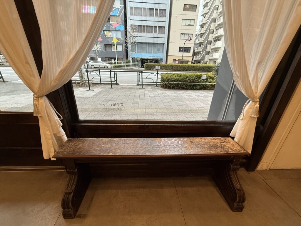 Antique-style wooden bench by the window inside Kashiya Shinonome with white curtains and street view