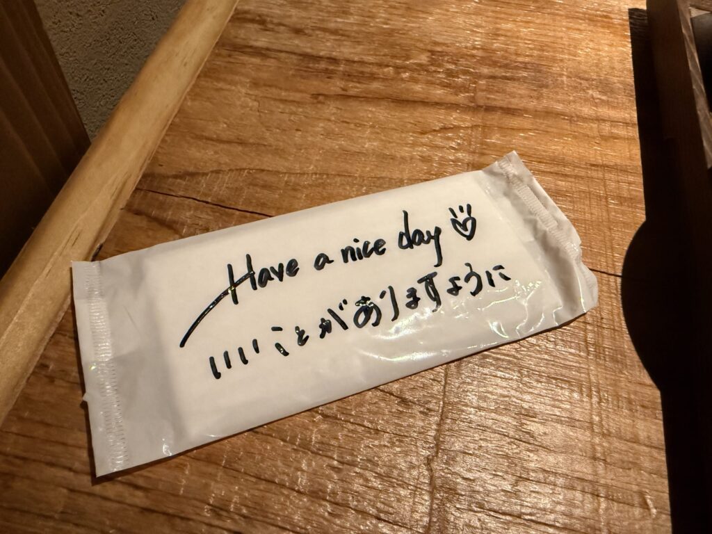 Oshibori wet wipe package on a wooden table with a handwritten message saying 'Have a nice day' and 'May good things happen to you' in Japanese.